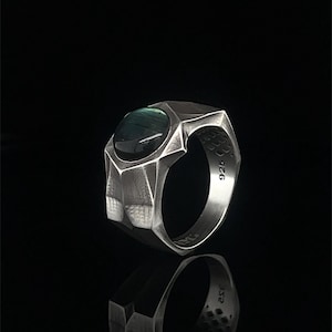 Puede incluir: Un anillo de plata con una banda geométrica facetada y una piedra ovalada, lisa y oscura engastada en el centro. El interior de la banda está claramente marcado con el número 925, indicando una composición de plata esterlina, sobre fondo negro.