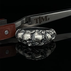 May include: A silver ring with three detailed skull designs. The skulls are intertwined with each other and have intricate details.