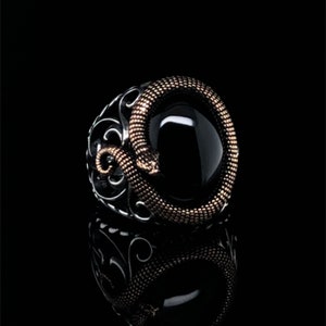 Peut inclure: Bague en argent avec une pierre d'onyx noire, ornée d'un serpent doré enroulé autour de la pierre. La bague présente des détails de filigrane complexes sur les côtés, créant un contraste saisissant.