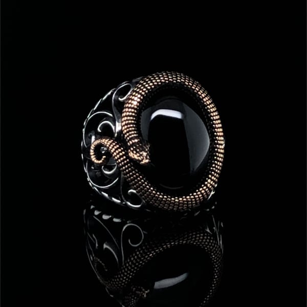 May include: A silver ring with a black onyx stone, featuring a gold-colored snake wrapped around the stone. The ring has intricate filigree detailing on the sides, creating a striking contrast against the dark background.