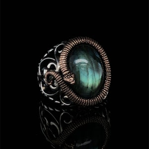 May include: A silver ring with a green gemstone set in a snake design. The snake is made of copper and has a textured surface.
