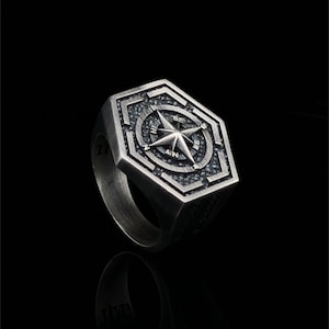 May include: A silver ring with a compass design. The compass features a star with points labeled N, S, E, and W. The ring is made of metal and has a textured surface.