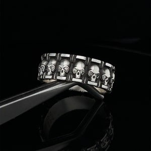 May include: A silver ring with a row of skull designs. Each skull has a detailed face with eye sockets and a prominent nose.