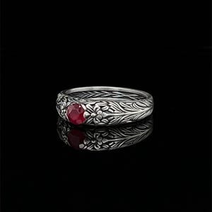 Könnte beinhalten: Ein silberner Ring mit einem runden, roten Edelstein in der Mitte. Das Ringband hat ein aufwendiges Blumen- und Blättermuster. Der Ring wird auf einer reflektierenden schwarzen Oberfläche gezeigt.
