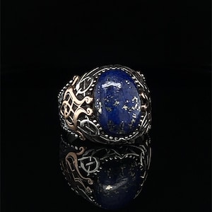 Puede incluir: Un anillo de plata con una piedra de lapislázuli azul engastada en un diseño ornamentado detallado. El anillo presenta un acento de oro rosa.