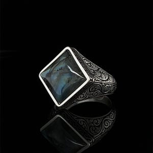 May include: A silver ring with a square, iridescent gemstone. The ring has an ornate, engraved design on the band. The gemstone has shades of blue, green, and gray. The ring is displayed on a reflective black surface.