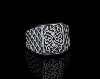 Op de afbeelding: Een zilveren ring met een vierkant ontwerp met een gedetailleerd, ingewikkeld patroon. De ring heeft een getextureerd, ruitvormig patroon aan de zijkanten.