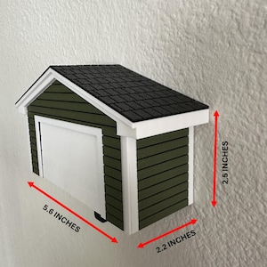 Op de afbeelding: Een miniatuur garagemodel, olijfgroen geschilderd met een witte deur en afwerking. Het dak is zwart en getextureerd. Afmetingen: 14,2 cm lang, 5,6 cm breed en 6,4 cm hoog.