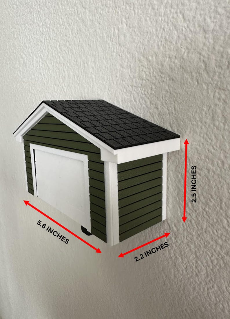 Op de afbeelding: Een miniatuur garagemodel, olijfgroen geschilderd met een witte deur en afwerking. Het dak is zwart en getextureerd. Afmetingen: 14,2 cm lang, 5,6 cm breed en 6,4 cm hoog.