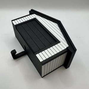 Op de afbeelding: Een zwart-witte decoratieve doos met een uniek ontwerp. De doos heeft een getextureerd oppervlak en een contrasterend patroon van zwarte en witte strepen. Er zit een haak aan de zijkant. De doos is ontworpen om rechtop te staan.