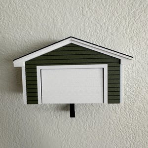 Op de afbeelding: Een miniatuur garagemodel met een witte garagedeur en een groen dak. Het dak heeft een witte rand en een zwarte omtrek. De garage is gemonteerd op een getextureerde witte muur.