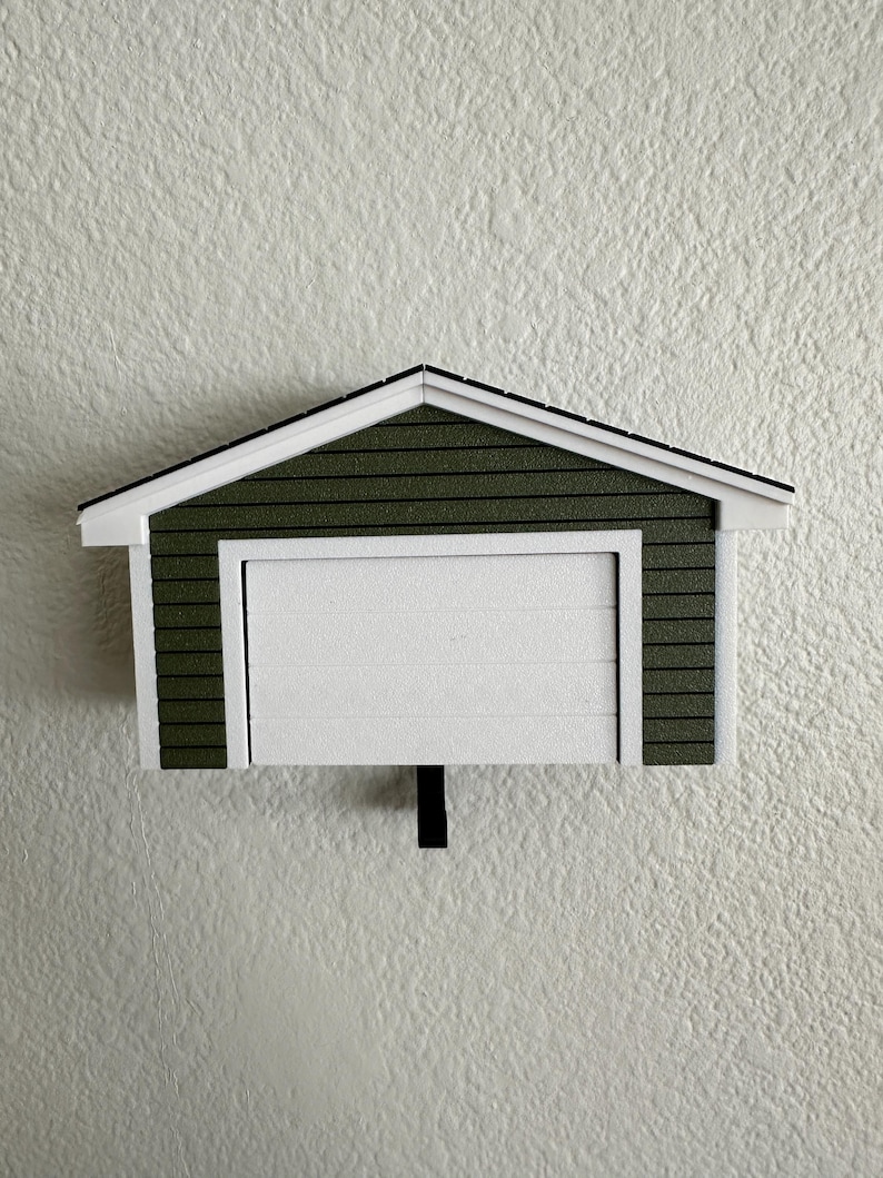 Op de afbeelding: Een miniatuur garagemodel met een witte garagedeur en een groen dak. Het dak heeft een witte rand en een zwarte omtrek. De garage is gemonteerd op een getextureerde witte muur.