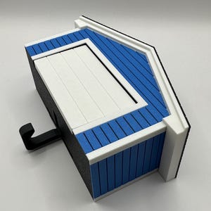 Op de afbeelding: Een miniatuur vogelhuisje in de vorm van een huis, blauw en wit, met een zwarte haak. Het dak is blauw met witte randen en de deur is wit. De zijkanten van het vogelhuisje zijn zwart.