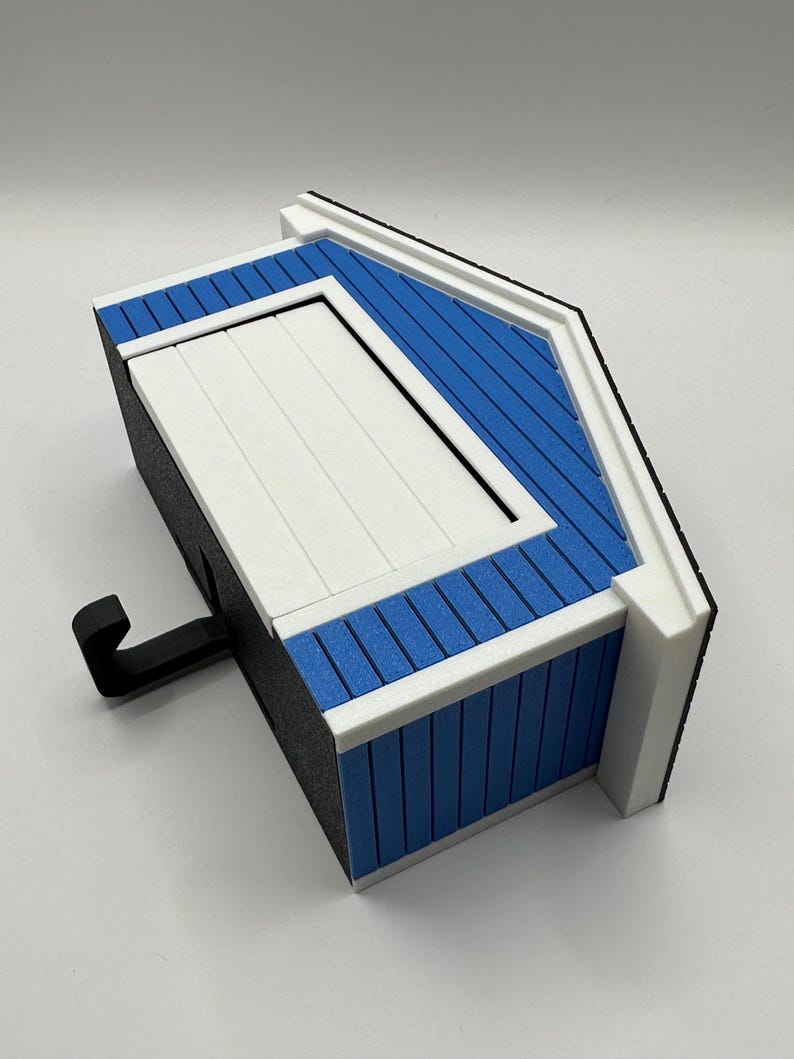 Op de afbeelding: Een miniatuur vogelhuisje in de vorm van een huis, blauw en wit, met een zwarte haak. Het dak is blauw met witte randen en de deur is wit. De zijkanten van het vogelhuisje zijn zwart.