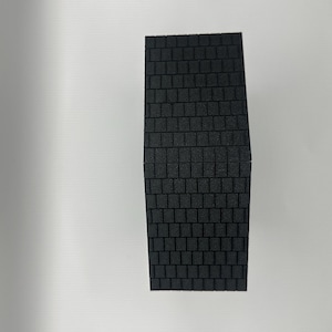 Op de afbeelding: Een rechthoekig, zwart schuimobject met een textuur, baksteenachtig patroon. Het object is donkergrijs van kleur en lijkt een bouwmateriaal of een modelcomponent te zijn. Het patroon is consistent over het oppervlak.