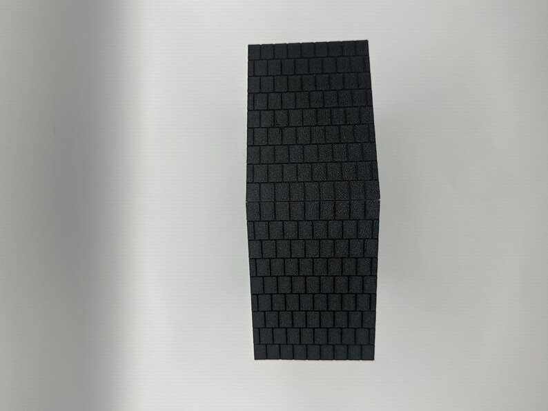 Op de afbeelding: Een rechthoekig, zwart schuimobject met een textuur, baksteenachtig patroon. Het object is donkergrijs van kleur en lijkt een bouwmateriaal of een modelcomponent te zijn. Het patroon is consistent over het oppervlak.