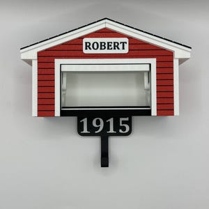 Op de afbeelding: Een rood-witte miniatuur garage met de naam "ROBERT" boven de garagedeur. Onder de garage staat een zwart bord met het nummer "1915". De garage heeft een zwart dak en is aan een witte muur bevestigd.