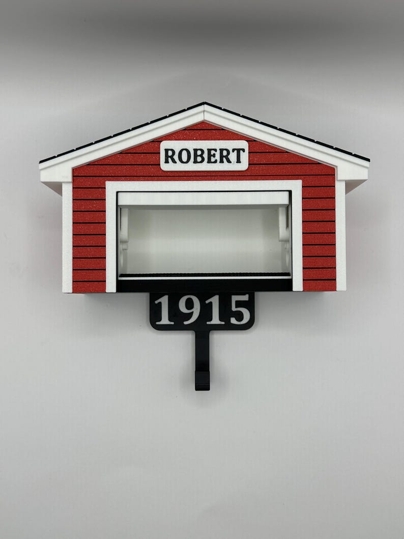 Op de afbeelding: Een rood-witte miniatuur garage met de naam "ROBERT" boven de garagedeur. Onder de garage staat een zwart bord met het nummer "1915". De garage heeft een zwart dak en is aan een witte muur bevestigd.