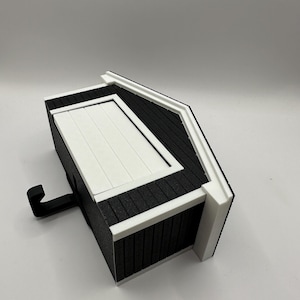Op de afbeelding: Een miniatuur zwart-wit decoratief schuurtje met een haak. Het schuurtje heeft een wit dak en witte randen, met zwarte gevelbekleding. De haak steekt uit de zijkant van het schuurtje.