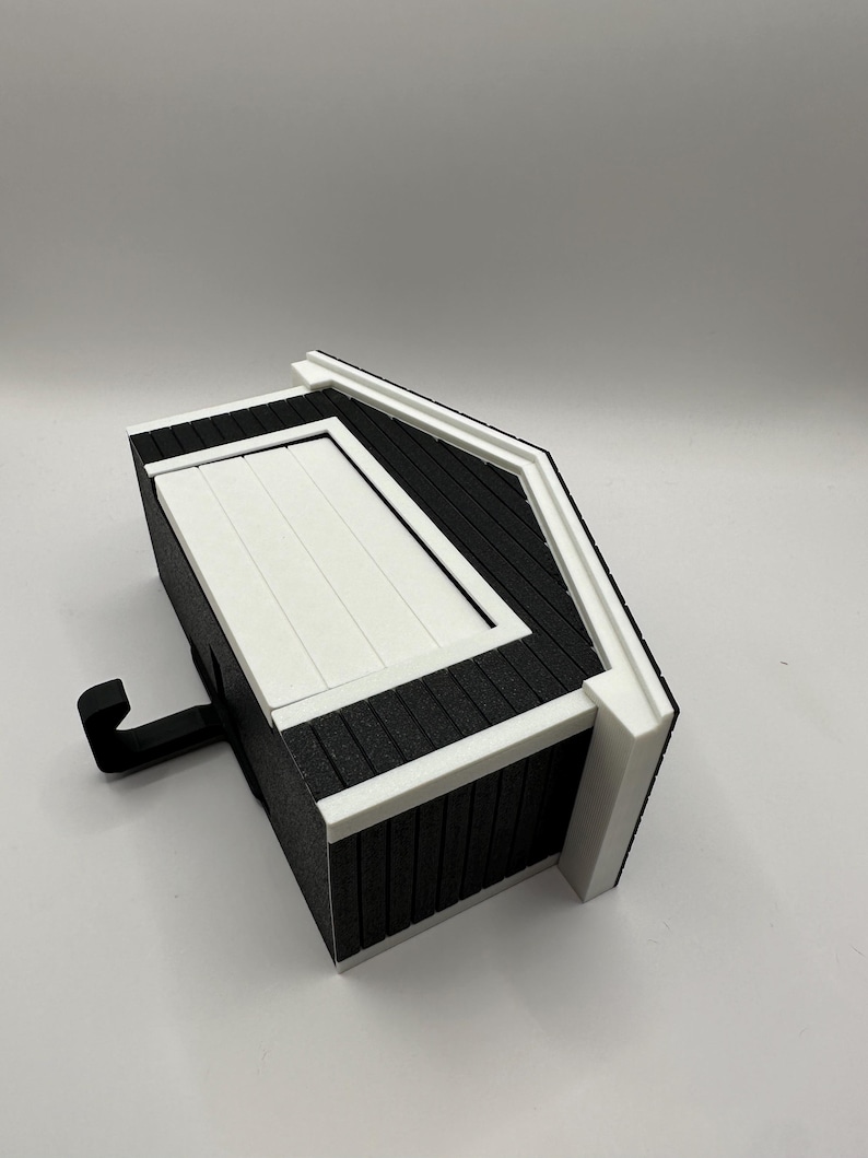 Op de afbeelding: Een miniatuur zwart-wit decoratief schuurtje met een haak. Het schuurtje heeft een wit dak en witte randen, met zwarte gevelbekleding. De haak steekt uit de zijkant van het schuurtje.