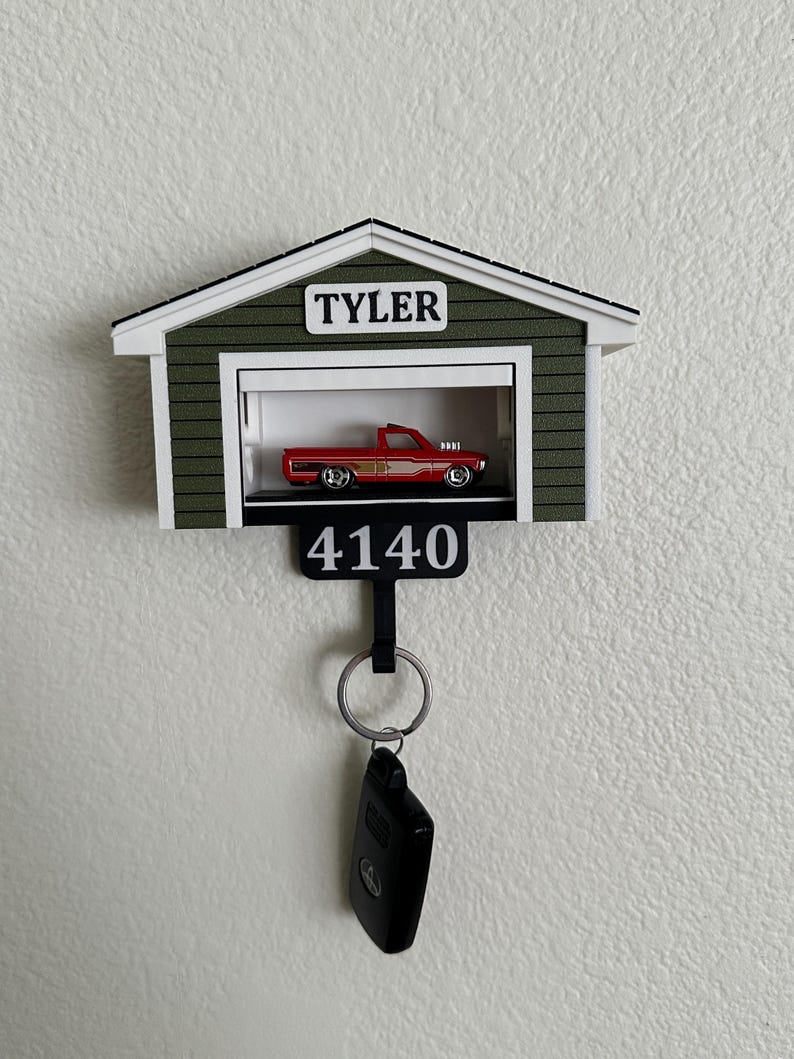 Op de afbeelding: Een decoratieve sleutelhouder in de vorm van een garage, met de naam "TYLER" boven de garagedeur. Binnenin staat een rode speelgoedtruck. Het nummer "4140" staat eronder, met een sleutelring en autosleutel.
