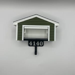 Op de afbeelding: Een huisvormige brievenbus met een groen en wit exterieur. De brievenbus heeft het nummer "4140" in zwart op een zwart bord met een haak eronder. Het dak is wit met zwarte rand.