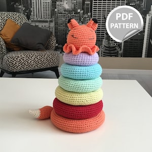 Puede incluir: Un juguete apilable de ganchillo hecho a mano con una cabeza de zorro sonriente sobre una pila de anillos coloridos. Los anillos son en tonos naranja, rojo, amarillo, verde, azul y morado. El zorro tiene una cola con punta blanca. La imagen incluye una pegatina "PDF PATTERN".