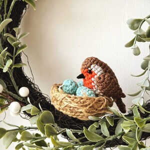 May include: A handcrafted wreath featuring a crocheted robin sitting in a nest with blue eggs. The wreath is adorned with green leaves, white berries, and a dark brown base, creating a charming spring or Easter decoration.