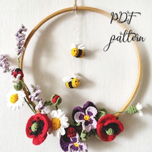 Könnte beinhalten: Ein runder Holzstickrahmen, verziert mit gehäkelten Blumen wie roten Mohnblumen, weißen Gänseblümchen und violetten Stiefmütterchen. Zwei kleine gehäkelte Bienen hängen oben am Rahmen. Der Text "PDF pattern" ist in der oberen rechten Ecke zu sehen.