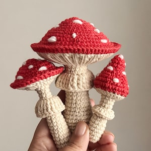 May include: A cluster of three crocheted mushrooms featuring bright red caps with white spots and cream-coloured stems. The central mushroom is the largest, flanked by two smaller mushrooms on either side, all crafted with visible yarn stitching.
