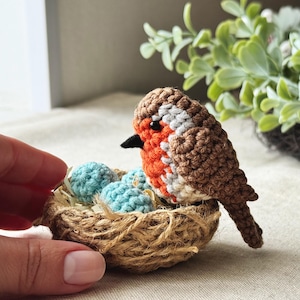 Op de afbeelding: Een handgemaakte gehaakte roodborst met een bruin lichaam, oranje borst en grijze en witte accenten. Het zit in een klein, geweven nest met drie blauwe eieren. De vogel heeft een zwarte snavel.
