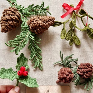 Op de afbeelding: Een verzameling gehaakte kerstversieringen, waaronder twee bruine dennenappels op groene ceder-takken, een takje maretak met een rode strik, een hulsttakje met drie rode bessen en een klein houten bordje met de tekst MEOLKA.