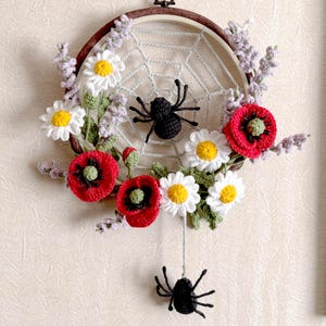 May include: A handcrafted dreamcatcher featuring a crocheted spiderweb, adorned with red poppies, white daisies, and lavender sprigs. Two black spiders are attached to the web. The dreamcatcher is set in a wooden embroidery hoop.