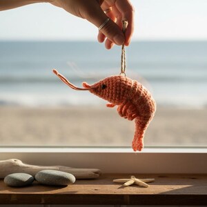 May include: A handmade, orange crochet shrimp ornament suspended by a string. The shrimp is held up by a hand, with a beach and ocean visible in the background. The shrimp has a detailed, textured appearance.