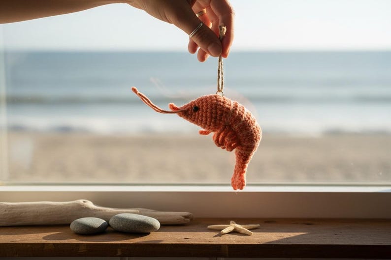 May include: A handmade, orange crochet shrimp ornament suspended by a string. The shrimp is held up by a hand, with a beach and ocean visible in the background. The shrimp has a detailed, textured appearance.