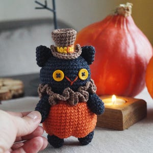 May include: A handmade crochet black cat doll wearing an orange outfit and a brown top hat. The cat has yellow button eyes and a brown ruffled collar. Two orange pumpkins and a lit candle are in the background.