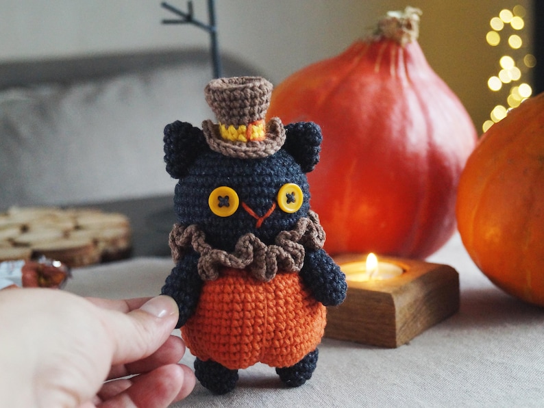 May include: A handmade crochet black cat doll wearing an orange outfit and a brown top hat. The cat has yellow button eyes and a brown ruffled collar. Two orange pumpkins and a lit candle are in the background.