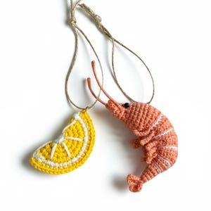 May include: Crocheted food ornaments: a yellow lemon slice with white details and a pink shrimp with white stripes. Both are suspended by twine. These handmade ornaments are perfect for kitchen decor or as unique gifts.