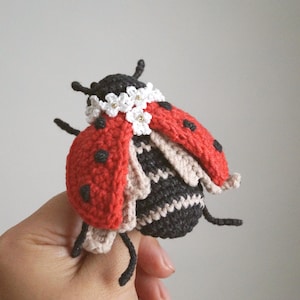 May include: A handcrafted crochet ladybird featuring a bright red shell with black spots, a black and beige striped body, and a decorative collar of small white flowers with yellow centres. The insect has black antennae and six thin black legs.