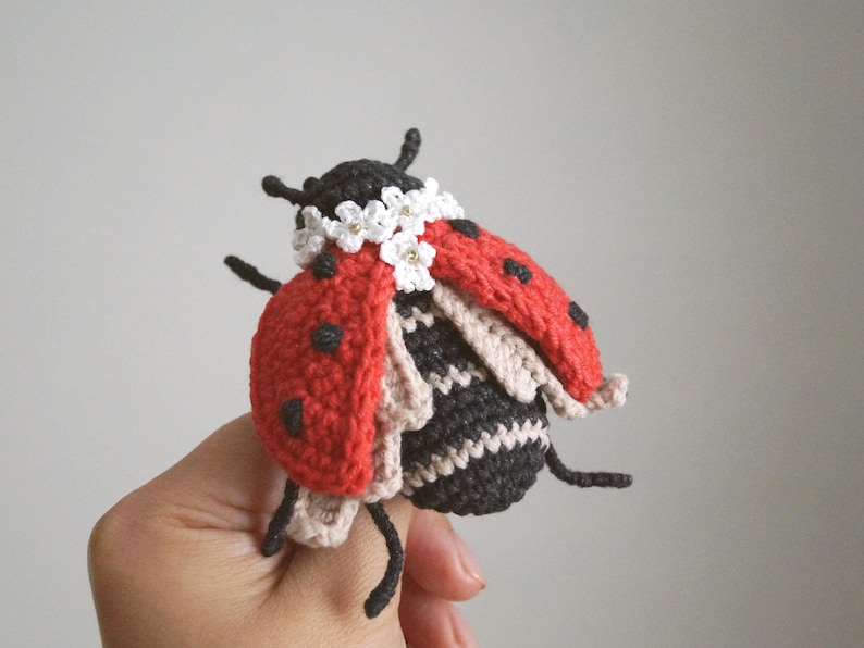 May include: A handcrafted crochet ladybird featuring a bright red shell with black spots, a black and beige striped body, and a decorative collar of small white flowers with yellow centres. The insect has black antennae and six thin black legs.
