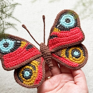 May include: A handcrafted crochet butterfly featuring vibrant red, mustard yellow, and bright blue circular patterns on its wings. The butterfly has a dark brown body and two thin antennae, with intricate black detailing surrounding the colorful eye-like spots.