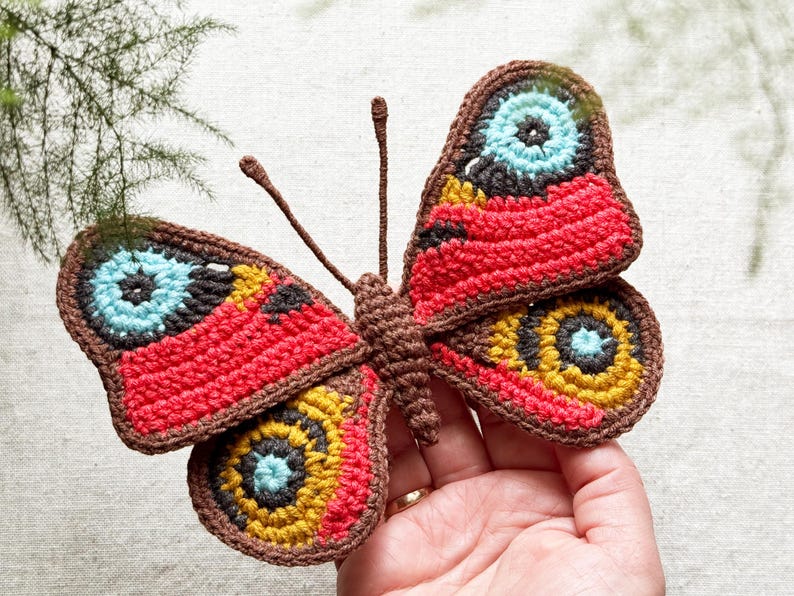 May include: A handcrafted crochet butterfly featuring vibrant red, mustard yellow, and bright blue circular patterns on its wings. The butterfly has a dark brown body and two thin antennae, with intricate black detailing surrounding the colorful eye-like spots.