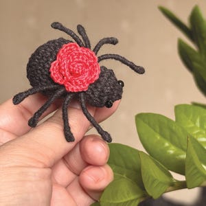May include: A small, dark grey crocheted spider featuring a bright red crocheted rose attached to its back. The spider has eight thin, dark grey legs and two small black bead eyes positioned on the front of its rounded, textured body.