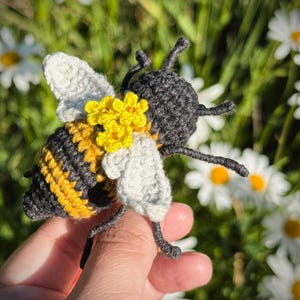 May include: A crocheted bee toy featuring a black and yellow striped body, light gray wings, and small yellow flowers attached to the back. The bee has dark gray antennae and legs, crafted with a textured yarn finish in a detailed amigurumi style.