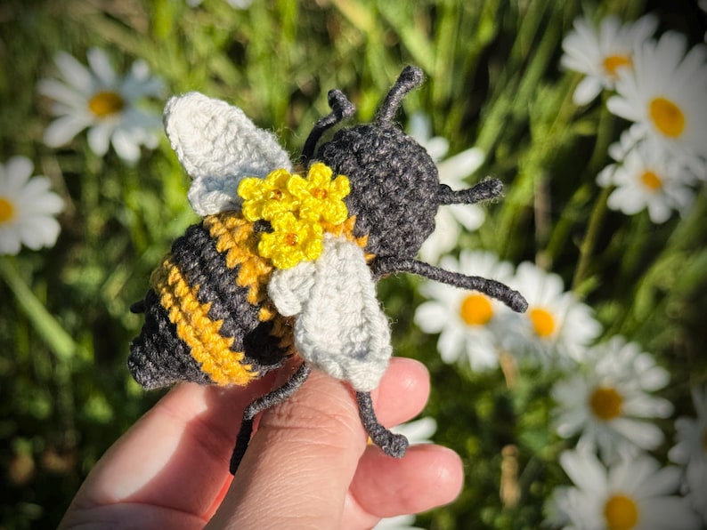 May include: A crocheted bee toy featuring a black and yellow striped body, light gray wings, and small yellow flowers attached to the back. The bee has dark gray antennae and legs, crafted with a textured yarn finish in a detailed amigurumi style.