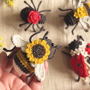 May include: A collection of four handcrafted crochet insects including a bee with a sunflower, a ladybug with white flowers, a spider with a red rose, and a bee with yellow flowers. Each insect features detailed stitching, dark bodies, and wings.
