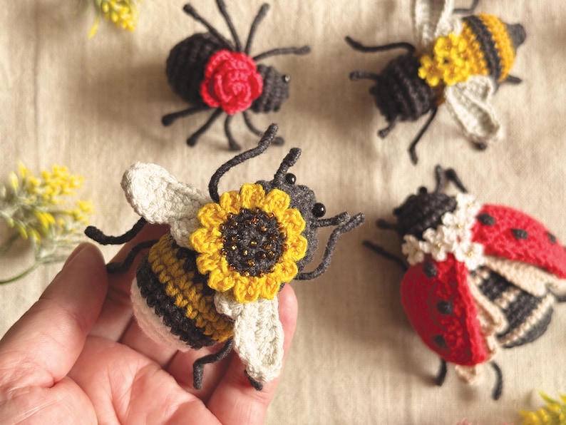 May include: A collection of four handcrafted crochet insects including a bee with a sunflower, a ladybug with white flowers, a spider with a red rose, and a bee with yellow flowers. Each insect features detailed stitching, dark bodies, and wings.