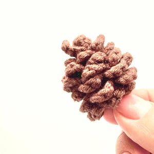 Op de afbeelding: Een bruine, gehaakte dennenappel, tegen een witte achtergrond. De dennenappel is gemaakt van getextureerd garen, met gedetailleerde ribbels en een natuurlijke vorm. Het knutselitem wordt in de hand van een persoon gehouden.