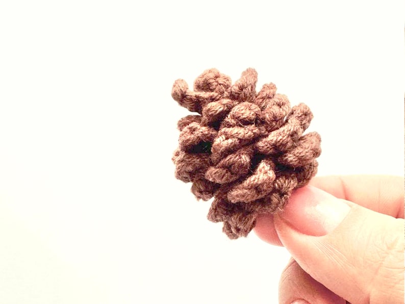 Op de afbeelding: Een bruine, gehaakte dennenappel, tegen een witte achtergrond. De dennenappel is gemaakt van getextureerd garen, met gedetailleerde ribbels en een natuurlijke vorm. Het knutselitem wordt in de hand van een persoon gehouden.