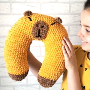 Op de afbeelding: Een geel gehaakt nekkussen in de vorm van een capibara. Het kussen heeft bruine accenten voor de oren, neus en voeten. Het kussen wordt door een persoon vastgehouden.